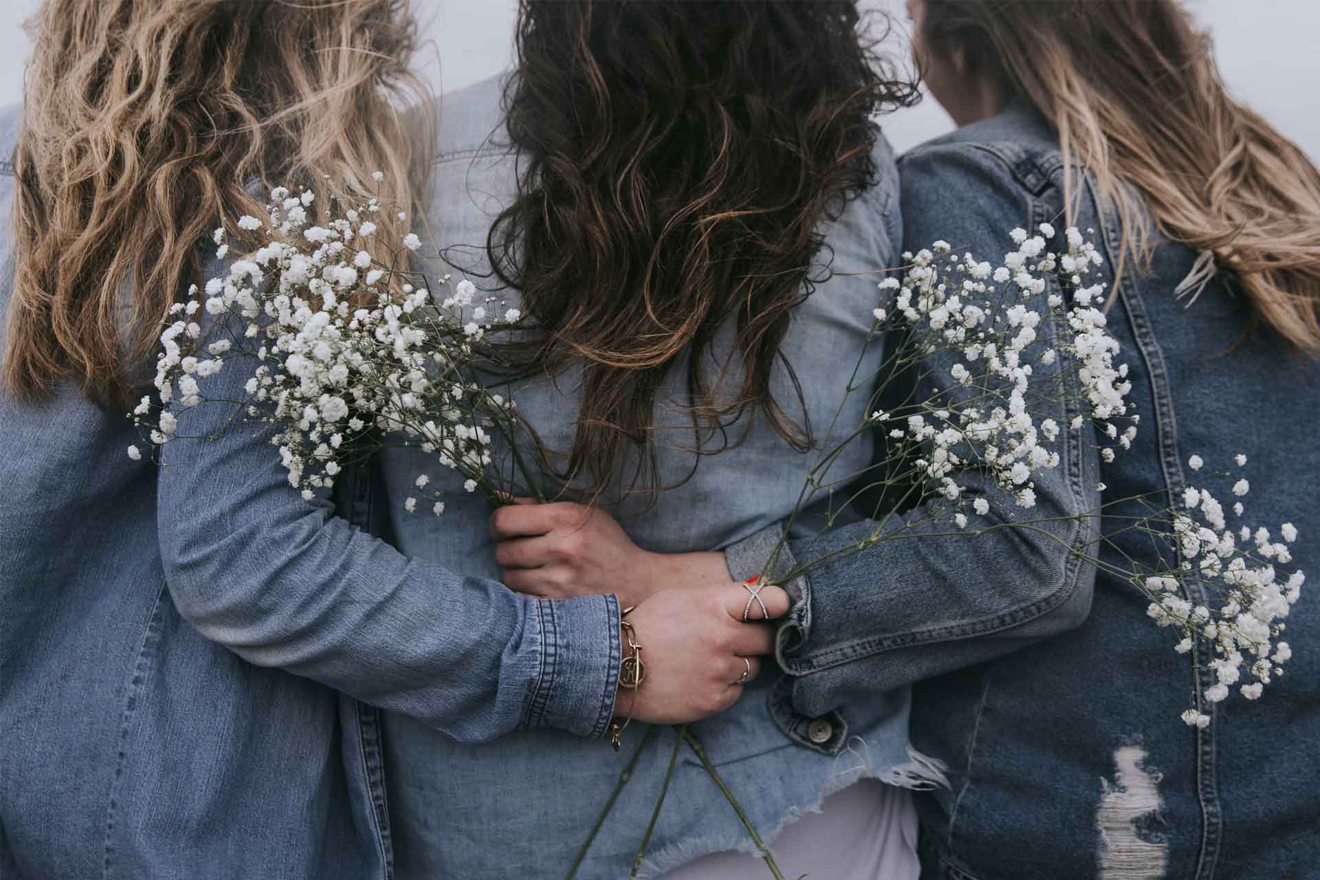 Tre donne viste di spalle si abbracciano affettuosamente, tutte e tre indossano giacche di jeans. Tengono in mano dei rametti di gypsophila (fiori bianchi). L'immagine trasmette un senso di amicizia, solidarietà femminile e calore, in un'atmosfera delicata e naturale.